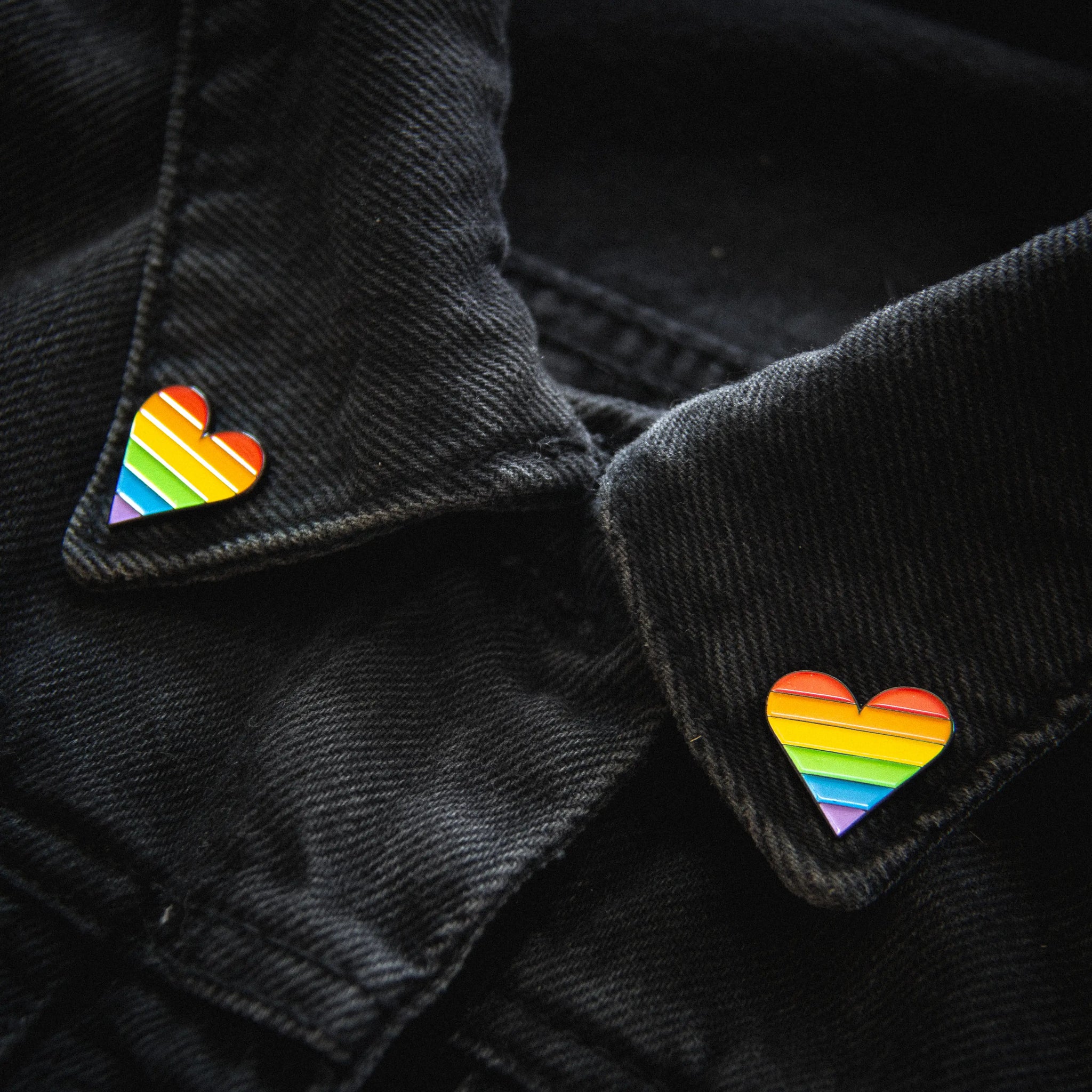 A set of rainbow heart shaped enamel pins on the collar of a black denim jacket.