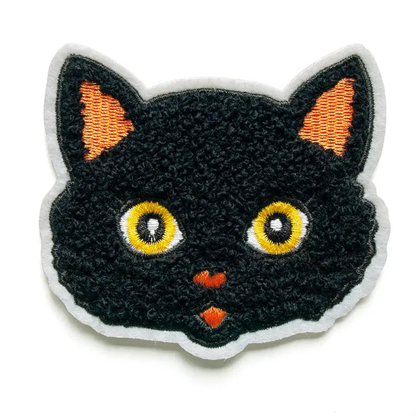 A patch of the head of a black cat in cartoon illustration style. The face is black chenille with embroidered orange ears, nose, and mouth. The cat’s eyes are yellow. The patch has a white felt border.