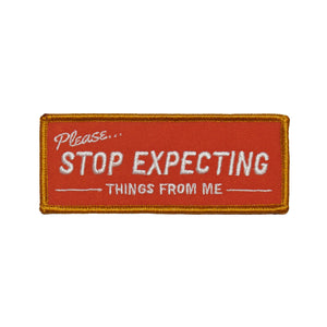 A rectangular embroidered patch with an orange border that is meant to look like a retro storefront sign. It reads “Please… Stop expecting things from me” in white. The background is a red-orange 