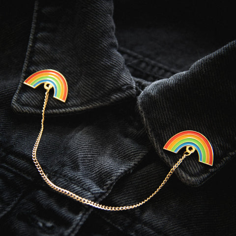 A set of two gold enameled pins of rainbows connected by a gold chain on the collar points of a black denim jacket 