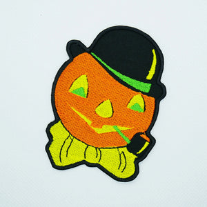 A cotton twill embroidered patch of a vintage Halloween decoration style carved pumpkin with a bowler hat, bow tie and corncob pipe