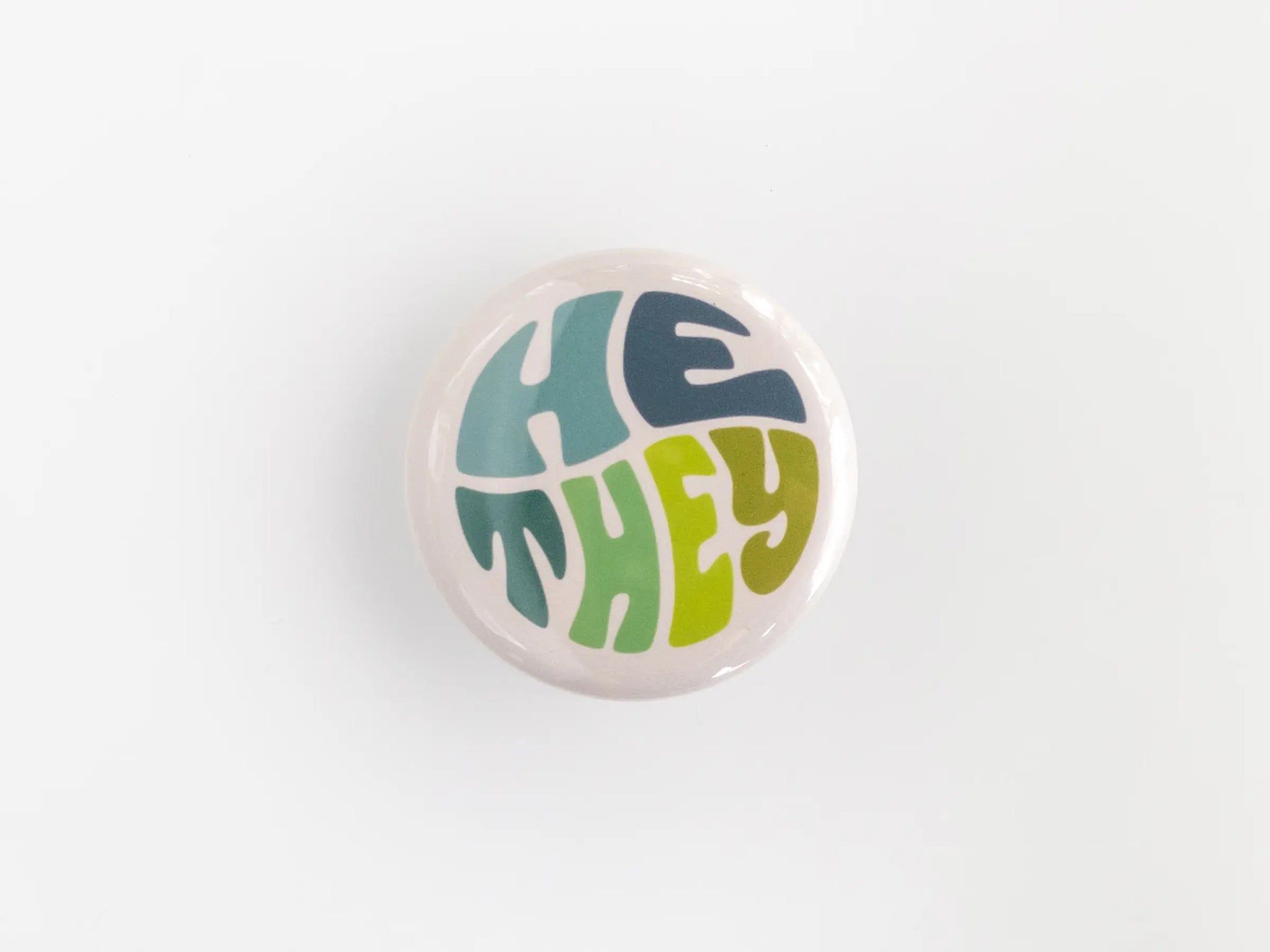 A 1.25” button with the pronouns “he” and “they” in multicolored lettering
