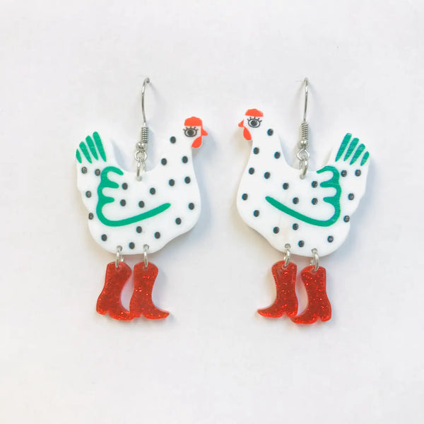 A pair of dangle earrings with silver fishhook hardware in the shape of a green and white chicken with black spots wearing a pair of red glitter cowboy boots. The boots are attached to the earrings with jump rings