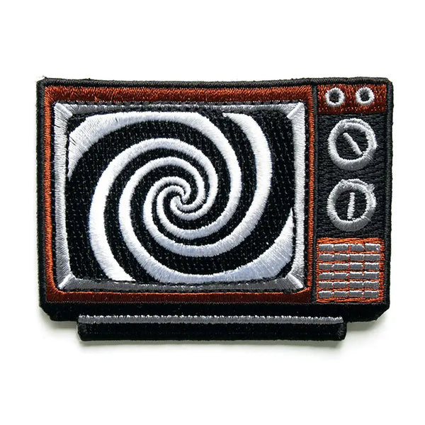 An embroidered patch of an old fashioned television set. It is brown with black and silver knobs and dials. On the screen there is a large black and white swirl