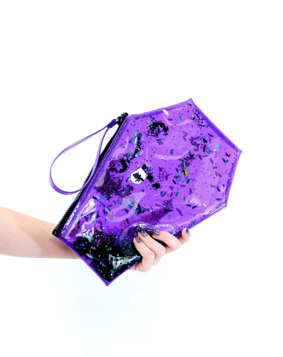 A hand holding a sparkly purple vinyl clutch purse in the shape of a coffin. It has liquid glitter inside of it in the shape of tombstones, bats, skulls, and stars
