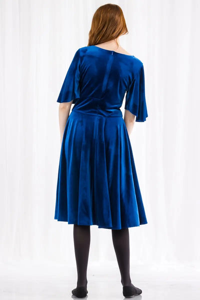 A royal blue velvet fit and flare dress with a deep plunging v sweetheart neckline, a wide banded waist, and elbow length flutter sleeves. Its skirt ends just past the knee. Shown on a model from behind to show back zip