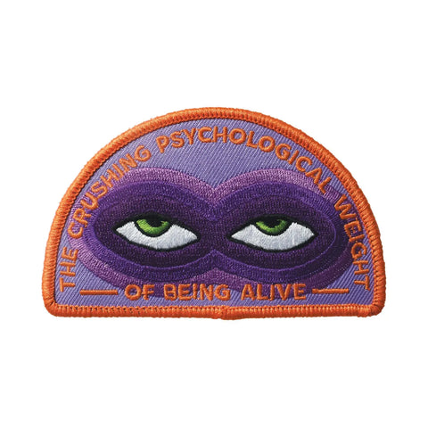 An embroidered patch with orange borders and writing saying “The Crushing Psychological Weight Of Being Alive”. There is a pair of green eyes surrounded by various shades of purple creating bags under the eyes 