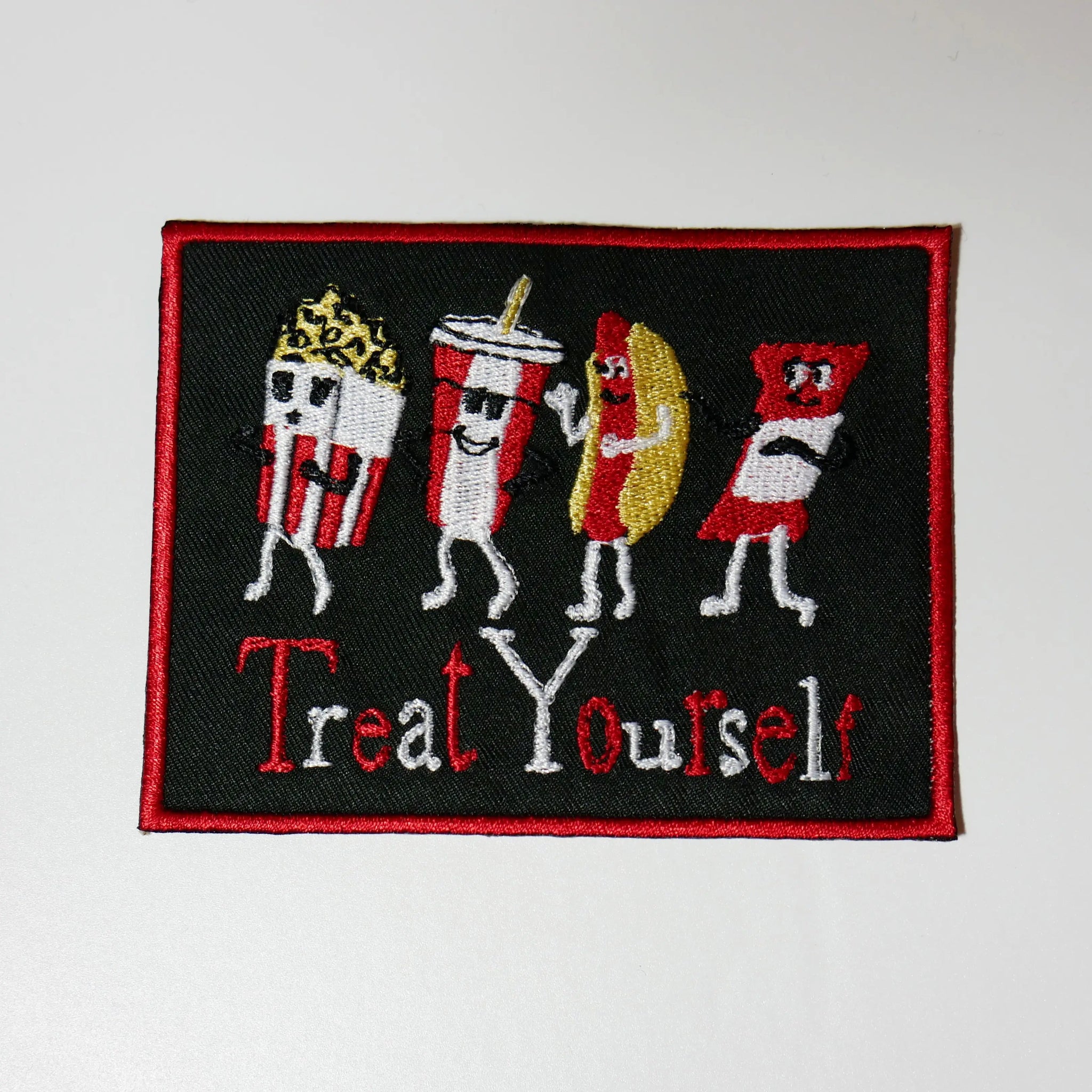 A rectangular patch depicting four classic movie theater treats with smiling faces- popcorn, soda, a hot dog, and a candy bar. Black canvas with a red border and lettering “Treat Yourself” in red and white 