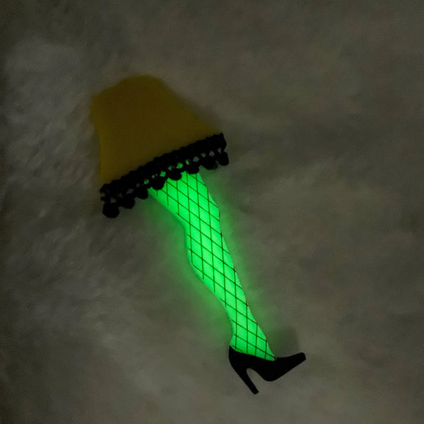 acrylic brooch of the iconic leg lamp from Bob Clark’s 1983 movie A Christmas Story with intricate hand-painted fishnet detail and pom-pom trim along the edge of the bright yellow lampshade. Shown in the dark to display the leg’s glow in the dark feature