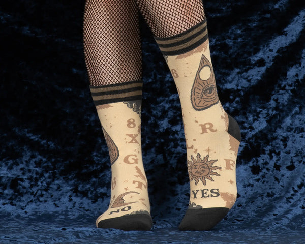 A pair of unisex crew socks with a spirit board design. Each pair has a beige background with dark brown heels and toes & a striped cuff of two shades of brown. Each sock has corresponding designs that when put together resemble the full markings of a spirit board including planchette. This shows a model wearing the socks overtop a pair of fishnets