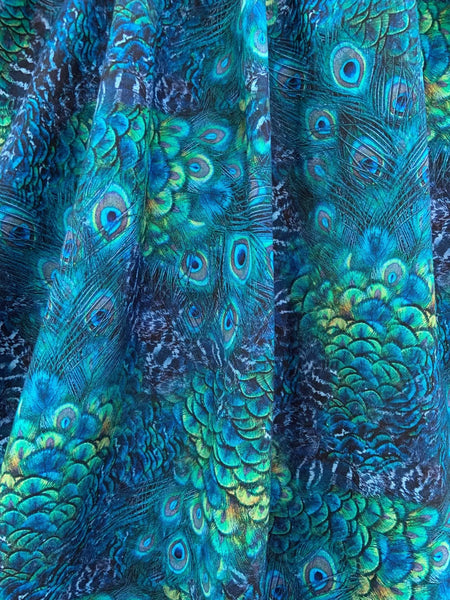 A sleeveless cotton dress in a green and blue peacock feather print with a surplus gathered neckline, wide gathered waist, and a just past the knee full skirt. A close up shot of the full skirt to show the pattern detail