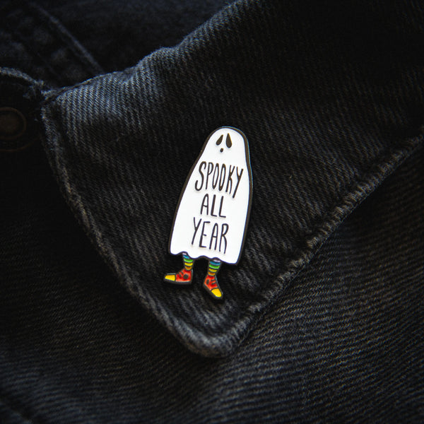 An enamel pin of a ghost wearing rainbow socks and red high top sneakers. The words “SPOOKY ALL YEAR” are written across its middle. The pin is on a denim jacket’s collar for scale