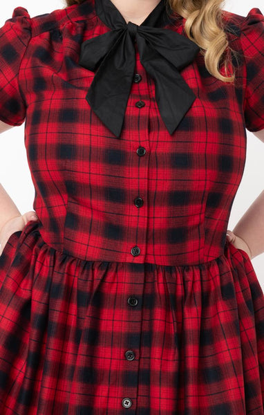 Cora Dress in rich red & black plaid features front button closure, fit & flare silhouette with gathered skirt in a just below the knee length, short puff shoulder sleeves, side seam pockets, and a black tie-closure collar. Shown cropped close up on model.