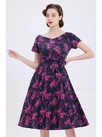 A model wearing a cocktail length short sleeved dress with full gathered skirt. It has a dark purple background patterned with black and pink reclining cats