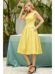 A model wearing a sleeveless dress in a shade of golden yellow with a fitted high neckline and princess seamed bodice, wide banded waist, full box-pleated just below the knee length skirt. Model is shown with one hand in one of the dress’ pockets
