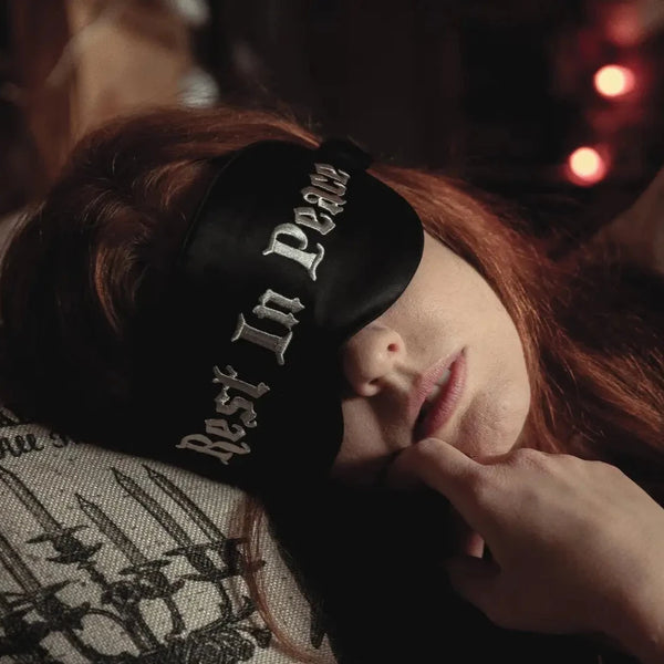 A model wearing a black silk sleep mask embroidered with “Rest In Peace” in white gothic font
