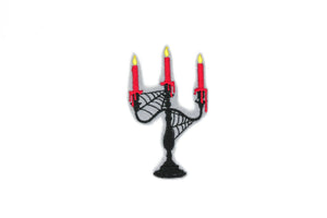 Embroidered patch of a black candelabra covered in spiderwebs holding three dripping red candles. On a light grey background 