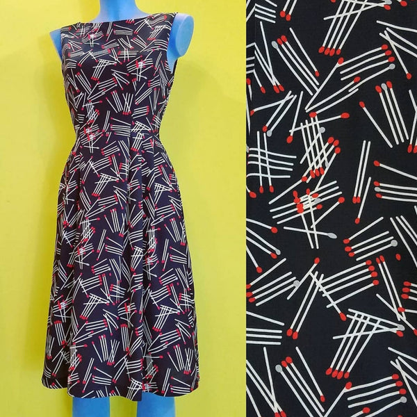 black with allover white & red stick matches print knee length sleeveless fit & flare dress, shown on mannequin next to fabric swatch close up