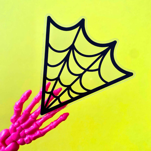 A die cut sticker of a black spiderweb on a clear background.  Shown being held by a skeleton hand to show clear background against a yellow backdrop