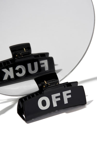 A black plastic claw style hair clip with “FUCK” printed on one side and “OFF” on the other side, shown reflected in a mirror