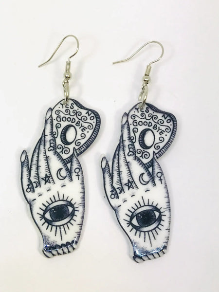 A pair of white acrylic coated earrings with an image of a hand covered in occult symbols with a large eye on its palm holding a ouija board planchette