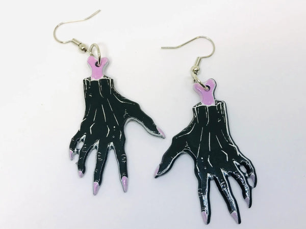 A pair of printed dangle earrings coated in acrylic resin- each earring has a severed hand with exposed pink bone and long pink fingernails 