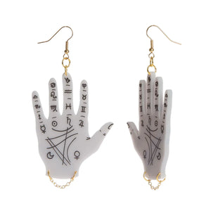 Dangle earrings of laser-cut light grey acrylic hands engraved and hand-painted with symbols used in the art of palm reading with a delicate gold metal chain hanging from the wrist of each hand