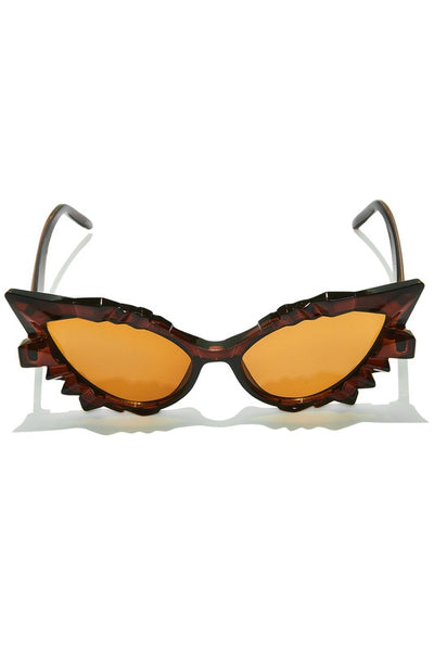 A pair of cat-eye sunglasses sitting at a front-facing angle. They are a translucent warm brown color and geometric detail above and below the lenses, which are brown