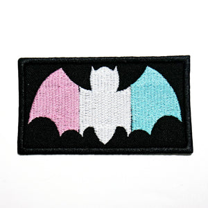 A black cotton twill patch with an embroidered bat in the pink, white, and blue colors of the trans pride flag