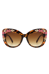 A pair of tortoiseshell rectangular cat eye sunglasses with pink and gold floral embellishments on the front of the frames
