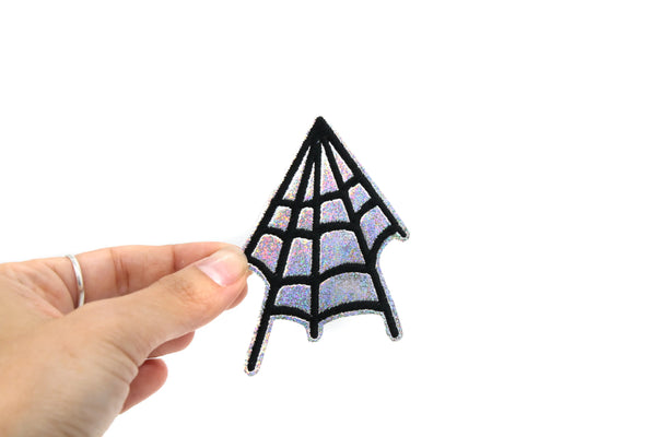 An embroidered patch of a black spiderweb on a silver holographic vinyl background. Held by a hand to show scale