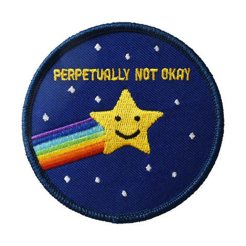 A round embroidered patch with a navy blue border. A metallic gold shooting star with a rainbow trail is in the middle of the patch surrounded by small white stars. At the top are the words “Perpetually not okay” in gold