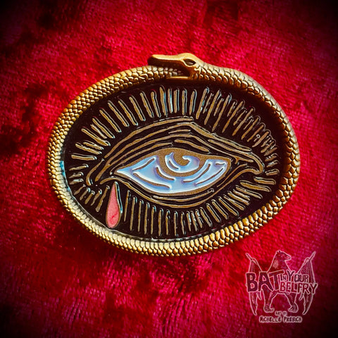 an antiqued gold metal pin of a weeping eye (illustrated in woodcut style) with a single iridescent red tear surrounded by a frame of a 3D sculpted ouroboros snake