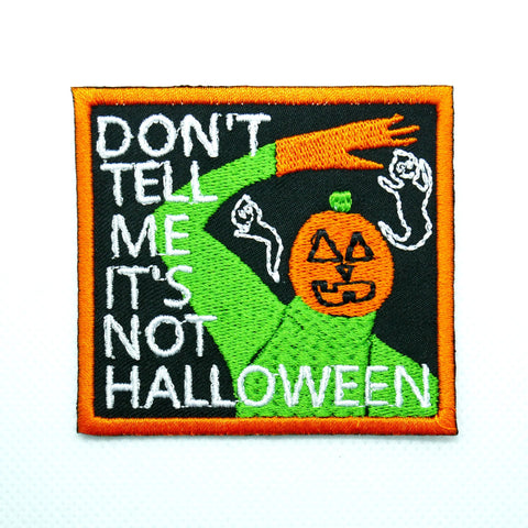 A rectangular black cotton twill embroidered patch with an orange border. It shows a creature with a green body and orange hands with a pumpkin head waving while surrounded by two small white ghosts. The words “Don’t tell me it’s not Halloween” is written in all capital letters down the left side of the patch