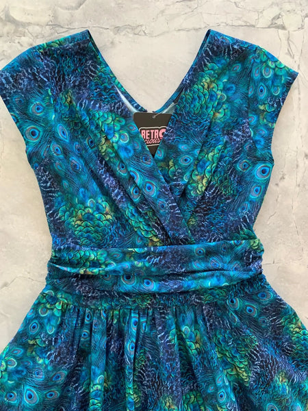 A sleeveless cotton dress in a green and blue peacock feather print with a surplus gathered neckline, wide gathered waist, and a just past the knee full skirt. Seen laid flat close up to show the gathered details on the neckline and waist 