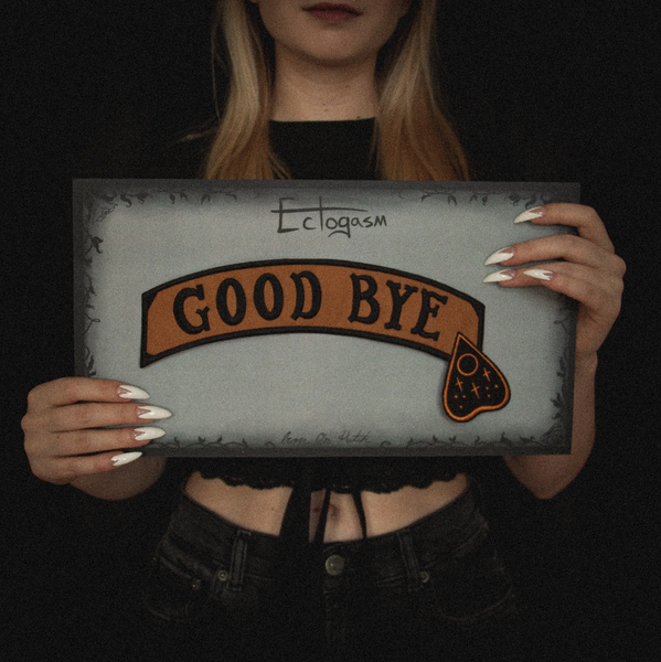 "GOOD BYE" message black stitching on light brown canvas banner with black Ouija board planchette embroidered back patch, shown on illustrated backer card packaging, held by model
