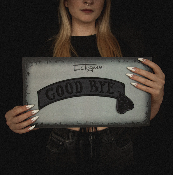 "GOOD BYE" message black stitching on dark grey canvas banner with black Ouija board planchette embroidered back patch, shown on illustrated backer card packaging held by model