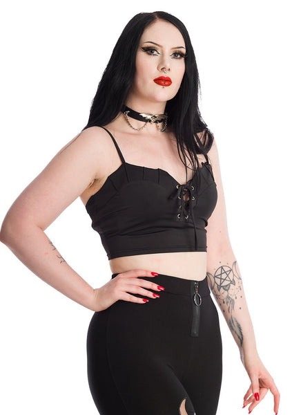 A model wearing a black bustier style crop top with a sweetheart neckline with batwing-style scalloped detail at the top of the bodice, 4 rows of lace-up eyelets with silver metal hardware, and adjustable spaghetti straps. Shown from the front at a three quarter angle to show bodice detail 