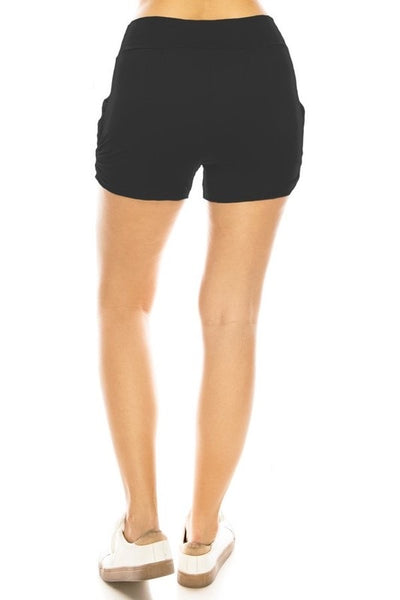 A model wearing black knit shorts with elastic waist band, pleated front, ruched side seam detail, and pockets. Seen from back