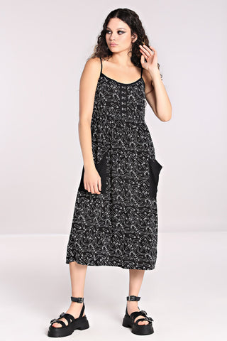 A model wearing a black sleeveless sundress with an all-over white safety pin pattern. The dress has black spaghetti straps and silver metal hook and eye detail at the bodice. The skirt is midi length with two black patch pockets at the hips.