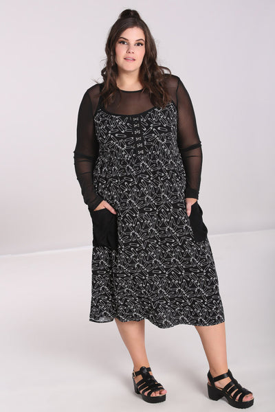 A plus size model wearing a black sleeveless sundress with an all-over white safety pin pattern. The dress has black spaghetti straps and silver metal hook and eye detail at the bodice. The skirt is midi length with two black patch pockets at the hips. The model is wearing a long sleeved mesh top underneath the dress and has their hands in the pockets of the dress