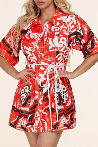 A model wearing a mini dress with a pointed collar and elbow length cuffed sleeves. The fabric is a swirly psychedelic pattern of black and white words on a bright red orange background. The dress has a single chest pocket and buttons down the front. It has full sized belt loops and a white cord style belt