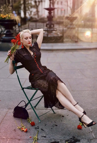 A model sitting wearing a black floral lace just past the knee dress. It has a v-neck shawl collar and cuffed short sleeves. There are bright red rose-shaped buttons running down the length of the bodice and on each patch pocket.
