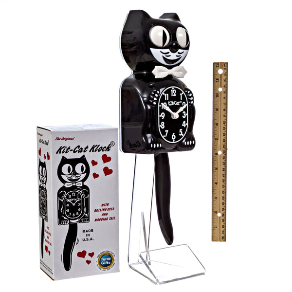black & white Kit-Cat wall mount clock features a mischievous grin, and big round eyes that swivel side-to-side in time with its pendulum tail , shown next to illustrated cardboard packaging and a ruler