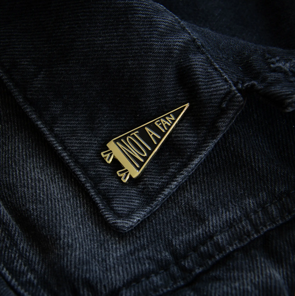 "NOT A FAN" message pennant shaped black enameled gold metal clutch-back pin, shown on denim jacket collar