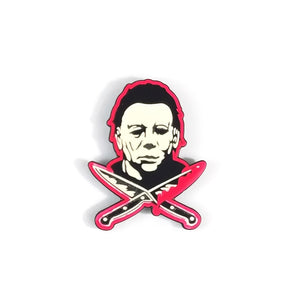 Enamel pin of Michael Myers from the Halloween movies surrounded by two knives in the style of a skull and crossbones, with a bright red border