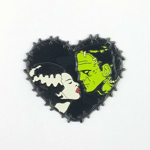 Glow-in-the-dark barbed wire trimmed black heart enamel pin depicting the iconic "We Belong Dead" profile poses of Boris Karloff as the monster and Elsa Lanchester as his mate from the classic 1935 movie, Bride of Frankenstein