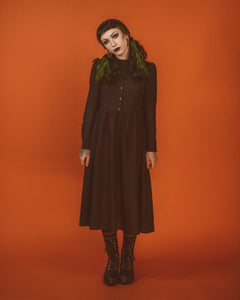 A model wearing a long sleeved tea length linen shirtwaist dress with slightly puffed shoulders and princess seaming on the bodice. It has small matte black buttons down the front. The full skirt is slightly gathered. The model is standing with their hands at their sides
