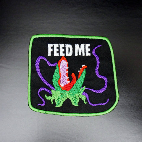 Audrey II from The Little Shop of Horrors in green, red, and purple under white "FEED ME" text embroidered on a green-bordered black canvas patch