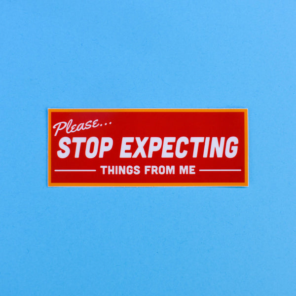 A rectangular vinyl sticker with an orange border that is meant to look like a retro storefront sign. It reads “Please… Stop expecting things from me” in white. The background is a red-orange 